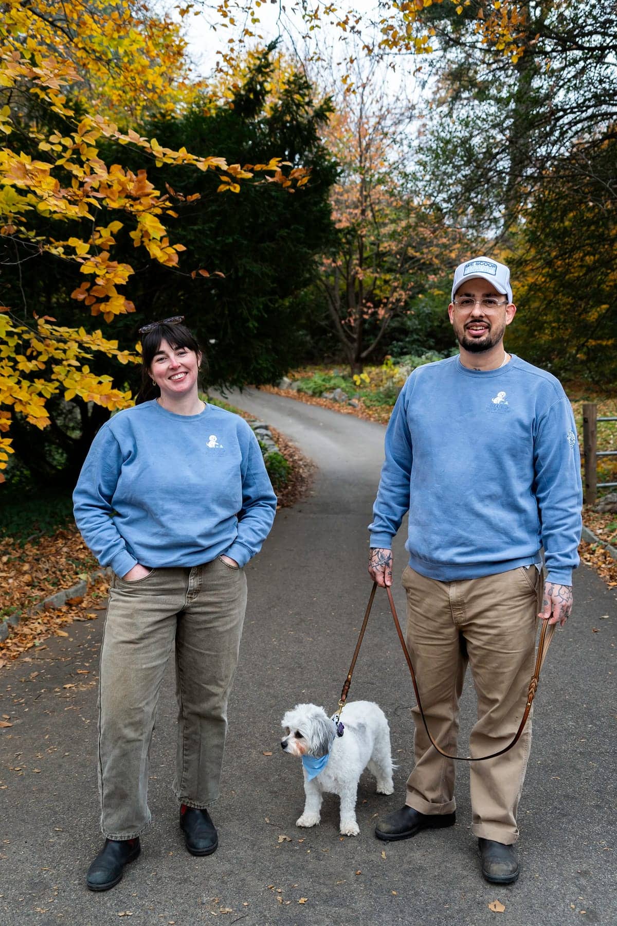 Both owners in branded sweatshirts on a leafy path, with their small white dog