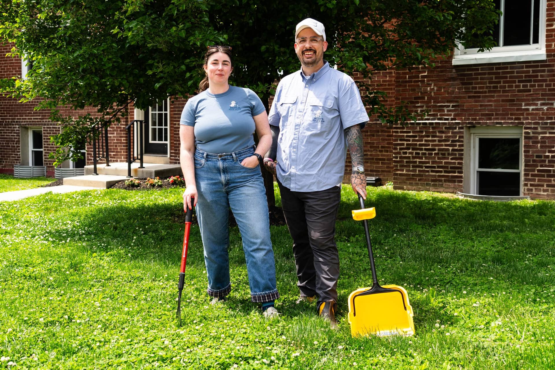 Two Blue's Poop Scoop owners on a lawn outside a brick building, holding a scooper