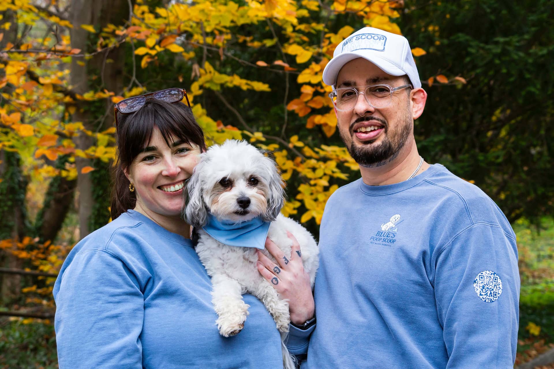 The owners of Blue's Poop Scoop in branded sweatshirts, holding their small white dog outdoors